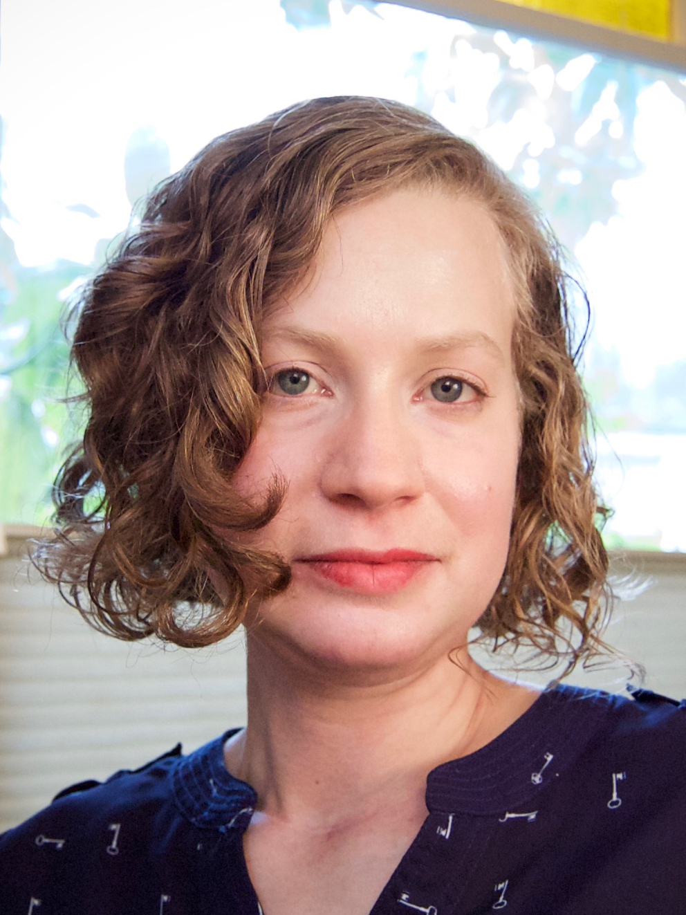 Anca L. Szilágyi looks head on at the camera, her curly, chin length hair framing her face. She is in front of a window and wears a dark shirt. 