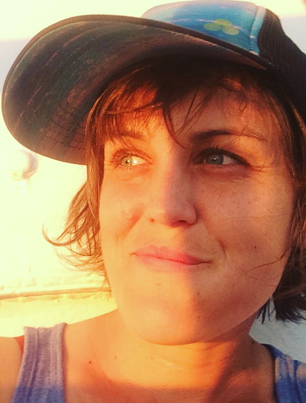 Catherine Edgerton half in sun, half in shadow. She smiles and looks out of frame to her right. She wears a tank top and a baseball cap, and has chin length brown hair with bangs.