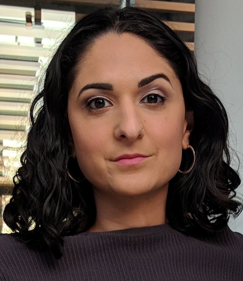 Sara Afshar smiles faintly at the camera with one dark eyebrow raised. She has shoulder length, wavy, black hair, warm eyes, and a dark shirt. 