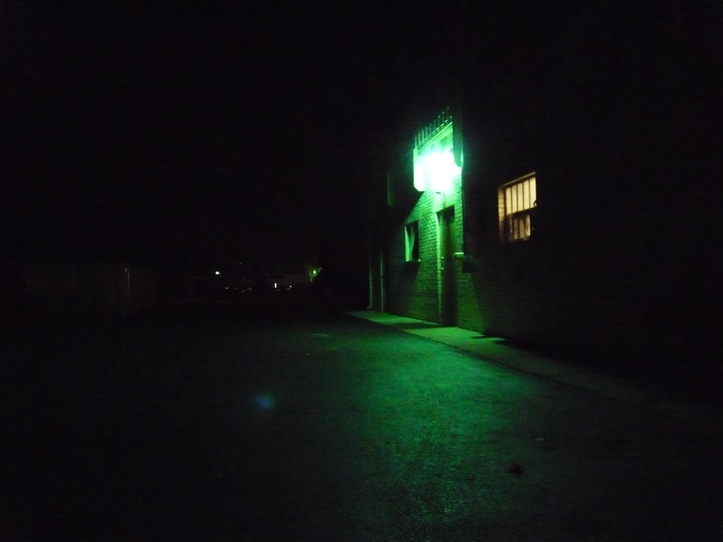 Green light over back door to brick building in dark street. Warm light, dimmer, comes through the window by the door. 