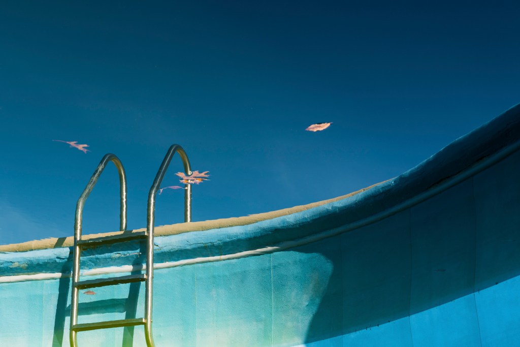 Ladder into a swimming pool, or perhaps out from a swimming pool. Everything is deep blue, bright at the bottom left, darkening at the top and to the right where the pool wall bends away. What looks like sky has three fallen leaves seemingly floating on the surface of water beyond where the ladder leads.