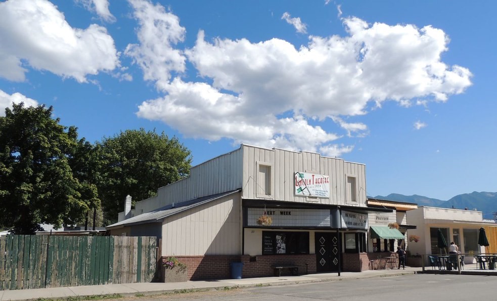Lincoln Theatre in Troy, Montana, A beige wooden building with a small marquee, under a blue sky with large fluffy clouds. Trees and mountains in the background.