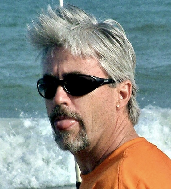 Hugh Findlay sticking out his tongue  at the beach. He wears sunglasses.
