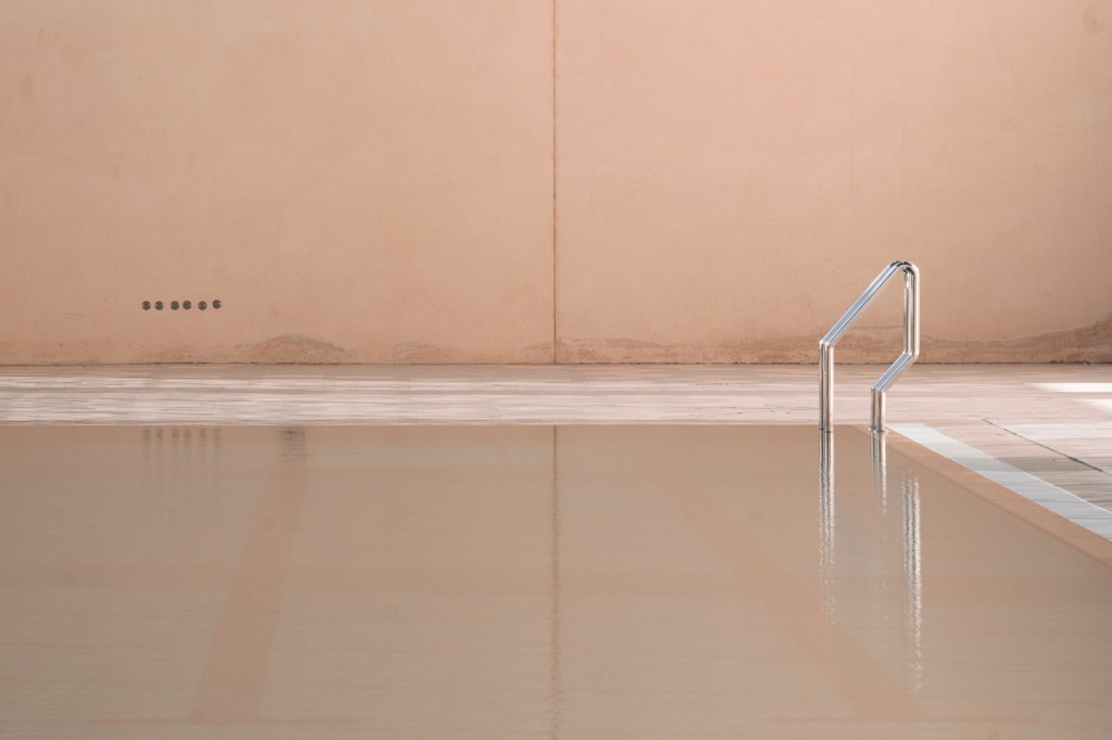 Pool in a peach room, pale tiles, metal ladder blurring and disappearing into water of unclear depth.