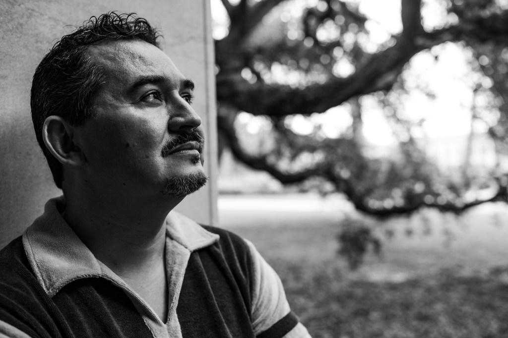 A black and white photo of Tomás Q. Morín looking off into the distance while leaning against a stone wall.