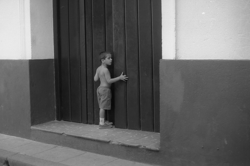 A black and white photo of a small boy standing outside on a step leading to a large dark wooden door. The boy has the door slightly opened and is peeking inside. 