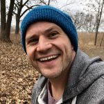 Christopher is wearing a blue beanie and a gray hoodie. He is smiling for the camera while walking outside. The trees are bare and all their leaves are on the ground.