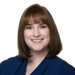 Laura has shoulder-length brown hair and bangs. Her hair is straight. She is wearing a navy blouse. The background is completely white.