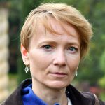 Veronica has light red hair, styled in a pixie cut. She is wearing small earrings. She is standing outside.