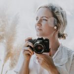 Evie is holding a camera and looking to the left. She has blonde hair tied low. She is wearing a white blouse.