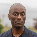 Eshun is a Black man standing outside. The background is blurry, but he appears to be in front of a body of water. He has no facial hair. He is wearing a navy sport coat.