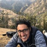 Madhav is smiling and kneeling down. He is in a hilly area with lots of trees. He has short black hair and glasses.