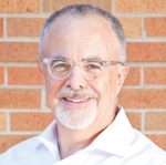 Gottlieb stands in front of a brick wall. He has very short grayish-white hair and a goatee. He is wearing glasses with clear frames.