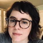 Jane has short bangs and shoulder length hair. She is wearing round glasses and a gray shirt.