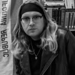 Black and white photo of White. He has long hair held back by a wide, black headband. He is wearing glasses and a leather jacket.