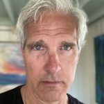 Roche looks into the camera, not smiling. He has white hair that falls over his forehead. He is wearing a black shirt.