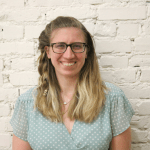 Eklund is standing in front of a white brick wall. She is wearing rectangular glasses. Her hair is down and she has one braid pulling her hair away from her face. She is wearing a light blue blouse. 
