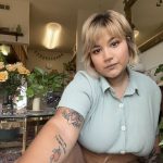 Pena is holding the camera to take a selfie. She is wearing a light-blue button up shirt, with short-sleeves. She has short blonde hair and bangs. Her tattoos are visible on her right arm. In the background, there are potted plants, flowers, and a bright ceiling light.