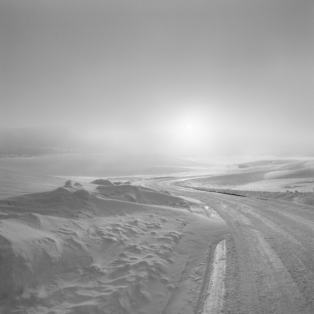 Black and white image titled Bozeman, Montana. The winding dirt roads fades out of view. Sand hills and a bright sun are the focus of this picture.
