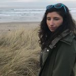 Marie is wearing a jacket standing on a beach. She is looking to the left, away from the camera. On the top of her long, dark wavy hair sits a pair of blue sunglasses.