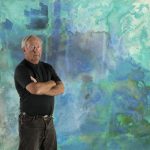 McPartlon stands in front of blue and green water color painting. He is wearing a black shirt tucked into black jeans. His arms are crossed. He has short white hair.