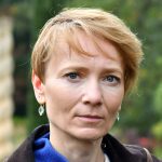 Winters has a pixie cut and light red hair. She is wearing earrings and looking straight into the camera with a very slight smile.