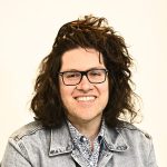 Rendoni has shoulder-length dark brown wavy hair. It is parted to the side. He has black rectangular glasses and is wearing a jean jacket. 