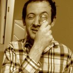 Joe Baumann squints and holds a small magnifying glass to his eye. He is wearing a plaid shirt and has short dark hair. The photo has a vintage-like yellow filter. 