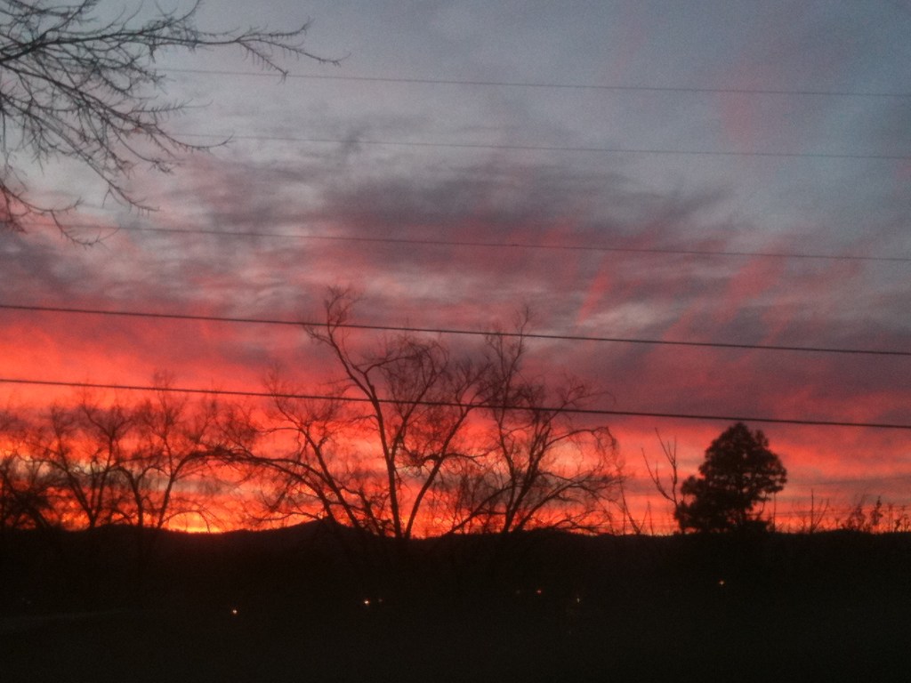 Untitled by Charles J. March III. A photo of the end of a sunset. The foreground of the image is almost completely black. The hills in the background stand out against the bright red orange sky. Five power lines cross through this photo. 