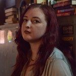 Nat has long magenta hair. Their body is turned away, but they are staring into the camera. They are wearing a long-sleeve satin blouse. 