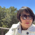 Photo of Margaret Juhae Lee, a Korean-American woman. She has short black hair and is wearing sunglasses. Her white coat has big buttons and a collar.