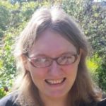 Photo of Gould from the shoulders ups. She has medium length blonde hair that is parted in the middle, tucked behind her ears. She wears small, rectangular glasses.