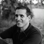 Rodrigo Toscano smiles in 3/4 profile, an arm thrown to the side, gazing past the camera. He's in black and white, outside somewhere.