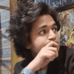 Khare is looking away from the camera. His hand is resting against his mouth. His hair is black, medium-length and has a lot of volume. 