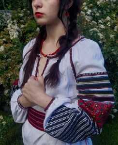 Resistance, by Anna Chornenka. This photograph is of a woman on a background of green bushes with bunches of white flowers. The image starts just below her eyes and extends to her hips. She has long braided brown hair, red lipstick, and clasps her hands over her heart. She wears a traditional embroidered white tunic with dark blue stripes and red flowers. 