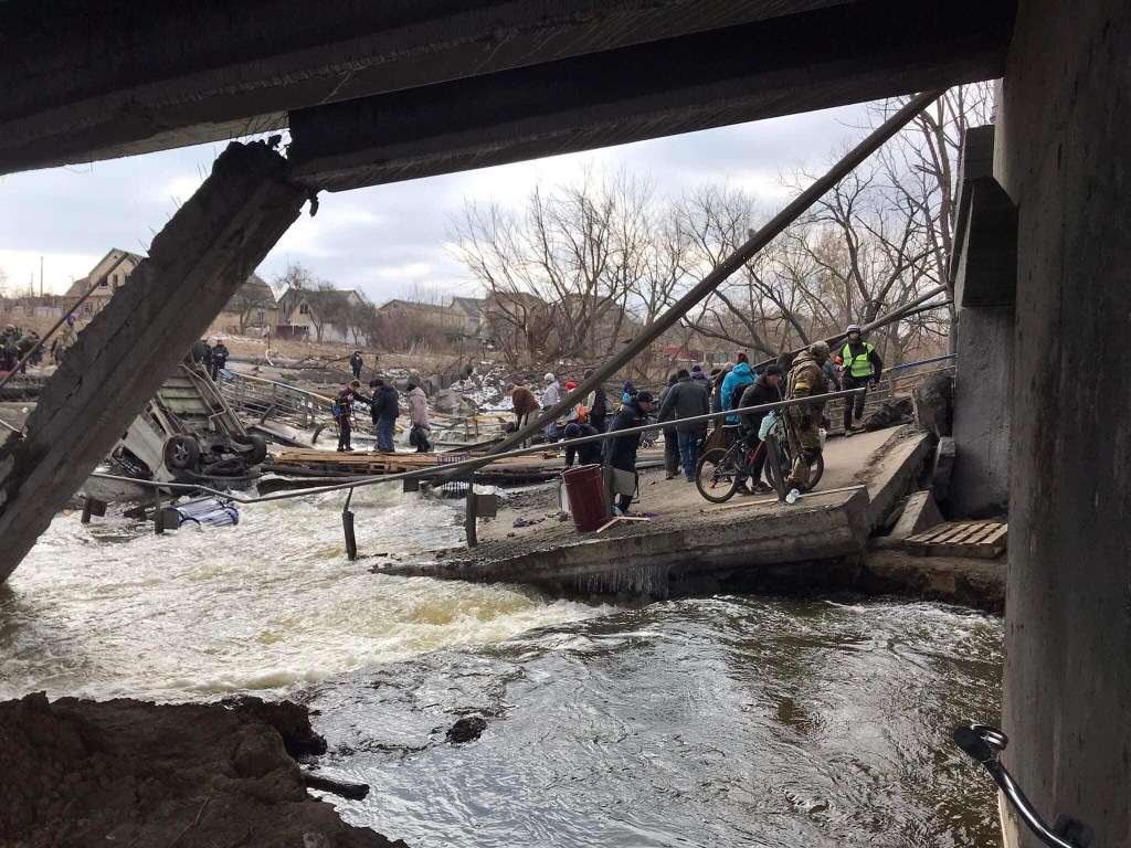 A photo of a group of people with bicycles crossing a river surrounded by debris from a collapsed bridge. The picture is from Victor G., and it is captioned "we put some lumber on the river so they could cross."