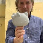 Mario Loprete holds a concrete Hello Kitty head out to the camera. He wears a blue button down, and has wavy grey hair that goes slightly past his ears. 