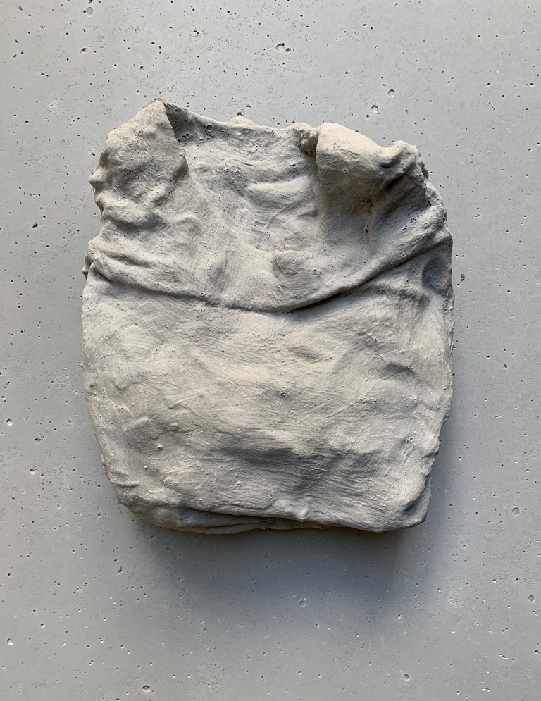 Untitled Concrete Sculpture 28 cm x 25 cm by Mario Loprete. This sculpture is concrete in the shape and texture of a folded shirt, fresh from the laundry. The neckline is at the top, with the sleeves hidden behind. The fabric appears to wrinkle slightly. The sculpture rests on a concrete backdrop, smooth, with small holes where bubbles once were. 