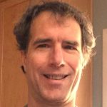 David Weaver has fluffy light brown hair. He smiles with his eyes as well as his mouth, in this close up of his face, lit slightly by the sun from his left. 