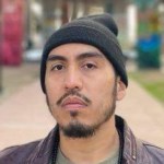 Esteban Ismael looks at the camera without smiling. He wears a dark beanie and a grayish purple jacket over a green shirt. He has a goatee and small mustache. 
