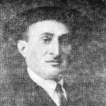Shloyme Gilbert, in a black and white photo distorted slightly by time. He wears a dark hat and smiles slightly. He has close knit eyebrows, a long nose and an expressive mouth. 
