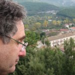 Alan Bern gazes down at a green cityscape, large buildings surrounded by trees. His face is in profile, he has fluffy graying brown hair and glasses. 