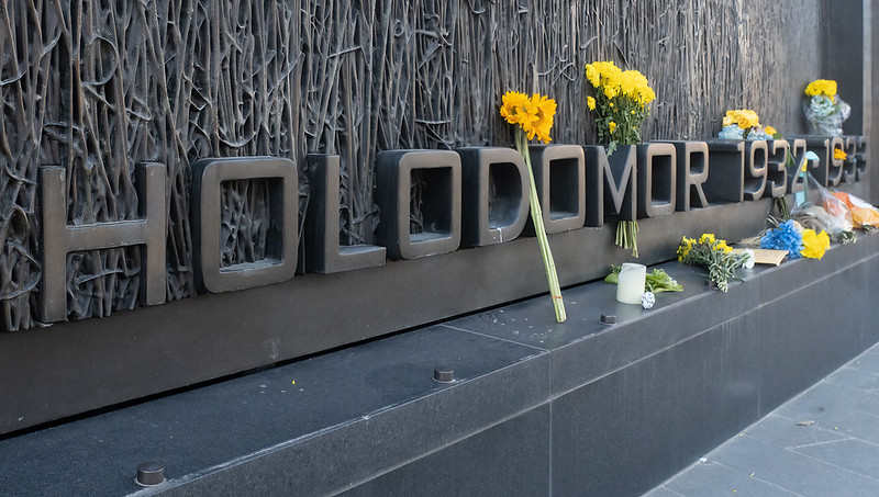 Holodomor Memorial in Washington DC, photographed in 2022 by Victoria Pickering. A wall of wheat behind the name Holodomor and the dates 1932-1933. People have left sunflowers, candles, bouquets and cards.