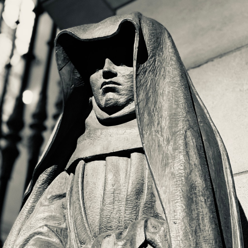 Hooded Nun by Jennifer Kircher Herman. A cream and black photograph of an ominous statue of a hooded nun, her eyes shadowed, her mouth frowning. She looms over the viewer, hands out of frame, all folded cloth and still stone. 
