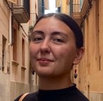 CC Molaison smiles in a narrow street between tall tan buildings with balconies and a sliver of blue sky. Her hair lies smooth against her head and her beaded earrings hang just above her black shirt. 