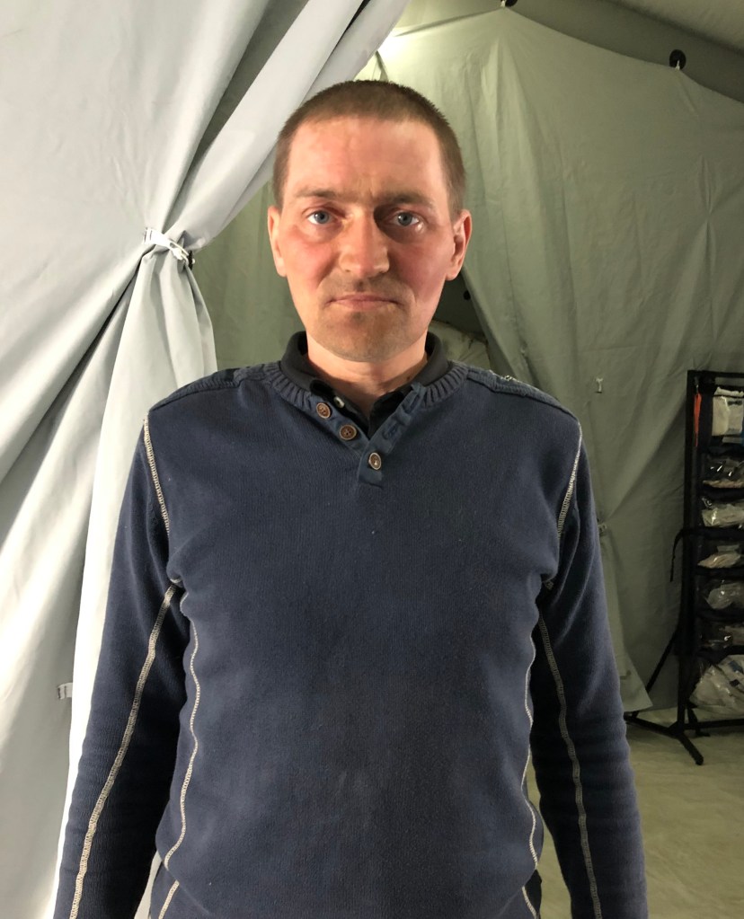Another untitled photo by Anna K. This is a man looking solemn with a buzz cut and a blue sweater. He stands in the entrance to a medical tent.