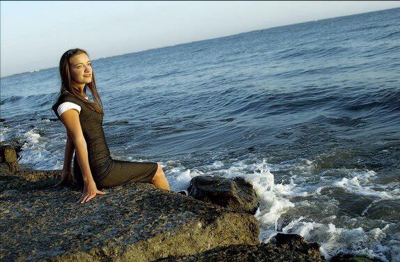 Laura Swart provides another photograph of the Sea of Azov, where a woman sits in a black dress with white sleeves, looking toward the sun across blue water, her legs dangling off a large stone. 