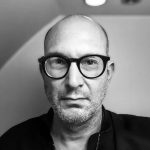 Interviewer photo: a black and white shot of Erik Noonan, who wears large rounded plastic frame glasses and a black shirt. He's in front of a corner where the ceiling bends to slope down, a lightbulb above his head.