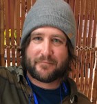 Edward Manzi wears a gray beanie and smiles slightly. His brown hair and short beard closely frame his face. He stands in front of a wood slat screen.