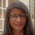 Reighan Gillam wears big plastic frame tortoiseshell glasses. Her long dark hair is parted on her right and she smiles at the camera, sitting in front of a white paneled window.